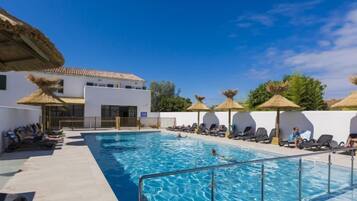 Une piscine entourée de chaises longues et de parasols, elle-même bordée d’un bâtiment blanc doté d’un toit en tuiles.