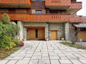 A multi-story building with a stone facade, wooden balconies, and a paved entrance area with potted plants.