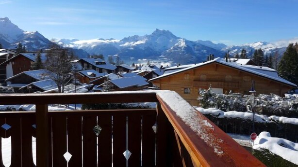 Un chalet in legno con un balcone affacciato su un villaggio montano innevato e un cielo azzurro limpido.