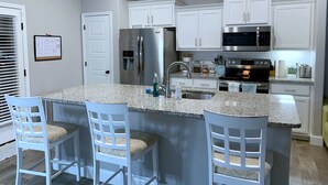 A modern kitchen with white cabinets, a stainless steel refrigerator, a microwave, and a granite countertop. There are three bar stools with patterned cushions. - MODERN HOME - firepit & fenced in backyard! (Carl Junction)