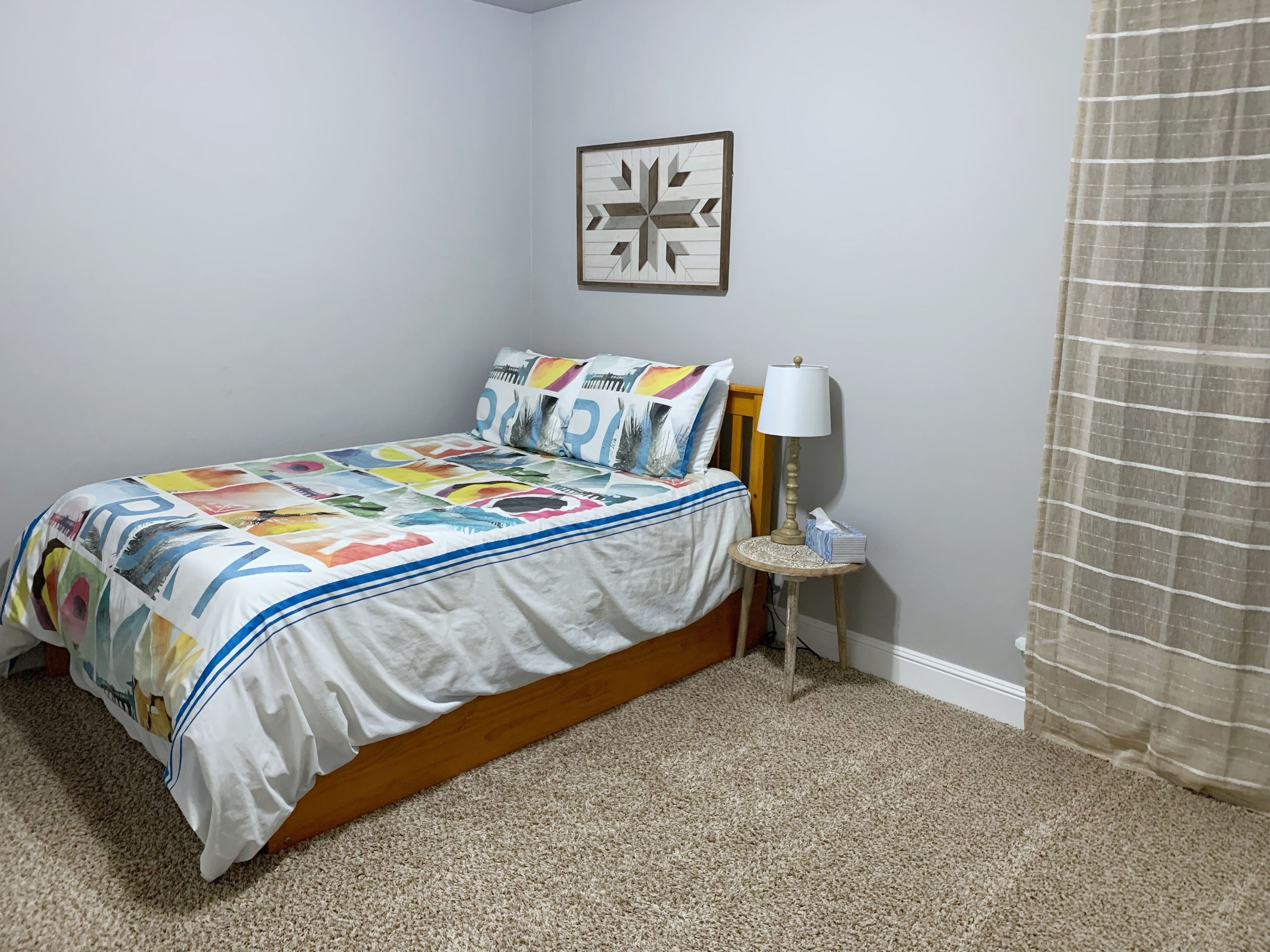 Een slaapkamer met een bed, nachtkastje met lamp, een lijst met foto aan de muur en een raam met gordijnen.