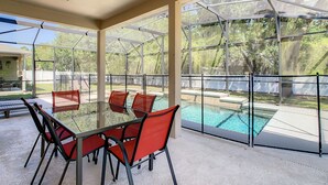 Uma área externa coberta com mesa de vidro e cadeiras vermelhas, com vista para uma piscina.