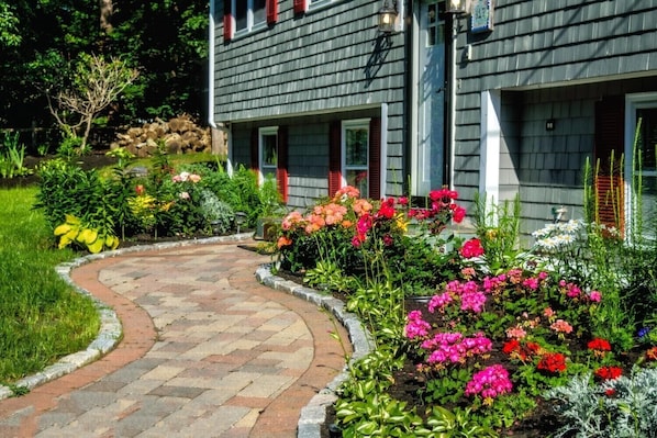 Une maison avec un jardin dont l’allée, pavée, mène à l’entrée, et qui est bordée de diverses plantes et arbustes en fleurs.