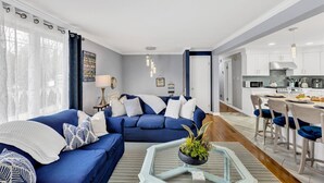 Uma sala de estar moderna com um sofá azul, uma mesa de centro de vidro e uma área de cozinha com armários brancos e uma mesa de jantar.