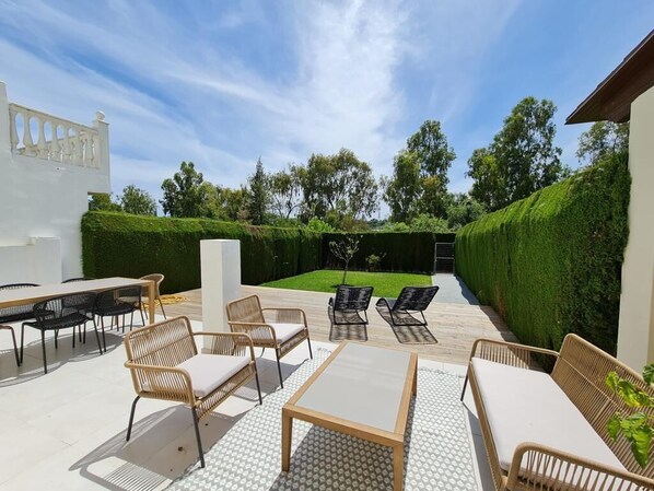An outdoor patio with wicker furniture, a table, and a view of a neatly trimmed hedge and trees in the background.