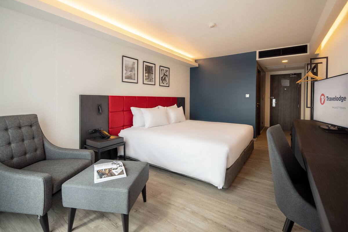 A hotel room with a bed, a grey armchair, a small table with a magazine, a desk with a computer, and a TV.