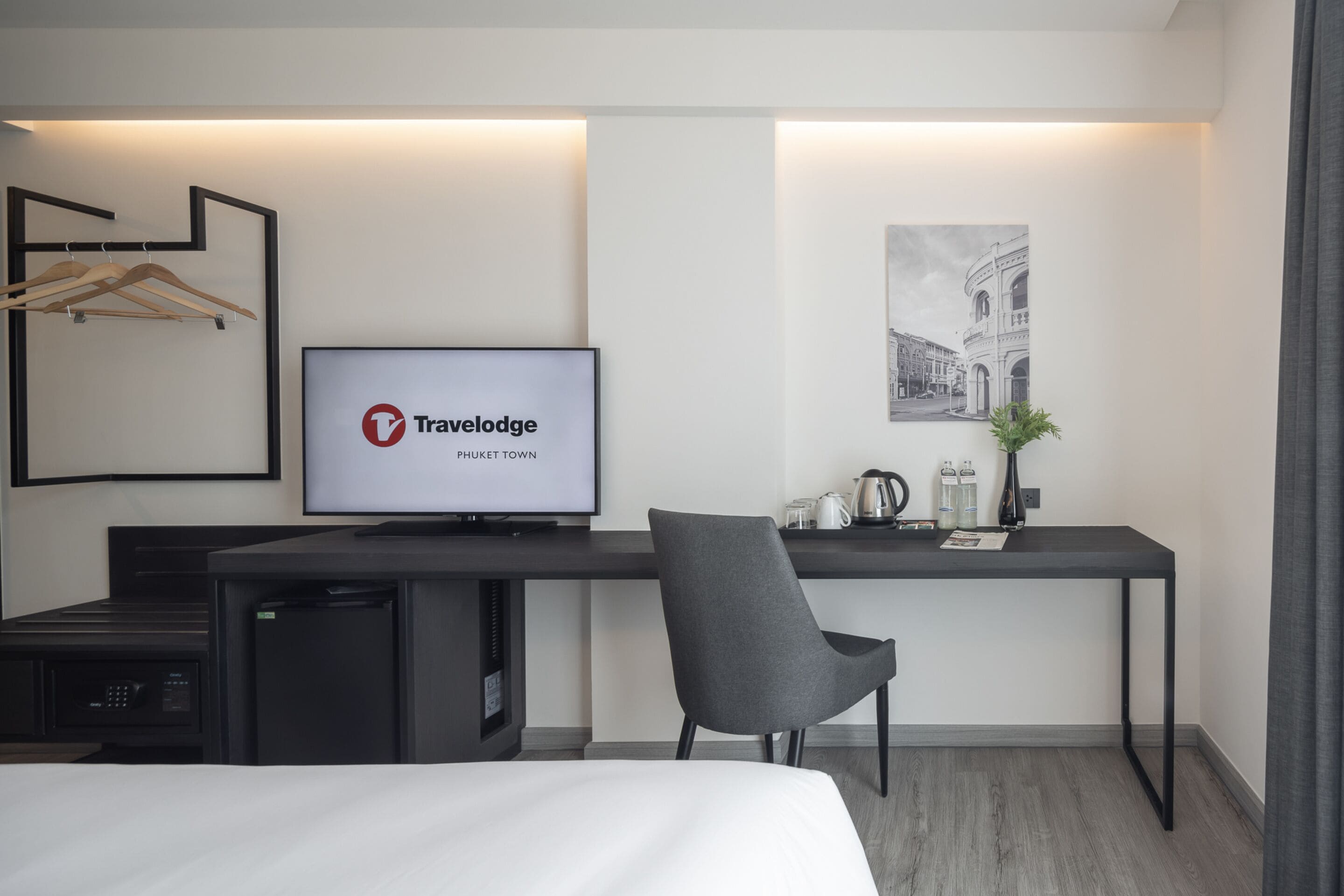Et moderne hotelværelse med en seng, et skrivebord med computer, en stol, et fjernsyn der viser 'Travelodge Phuket Town' og et billede i ramme på væggen.