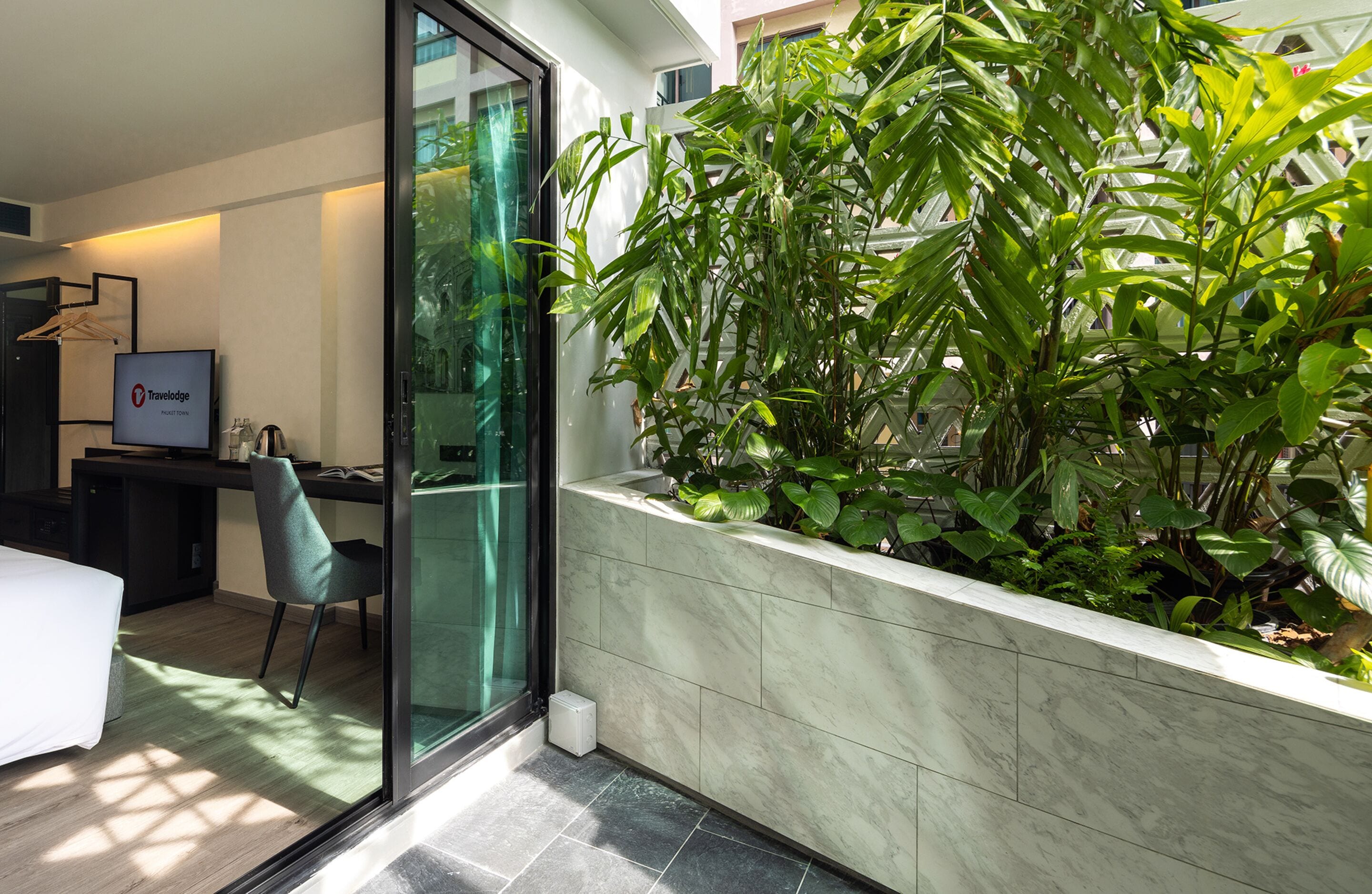 A modern hotel room with a glass door leading to a balcony, a desk with a computer, and a large potted plant.
