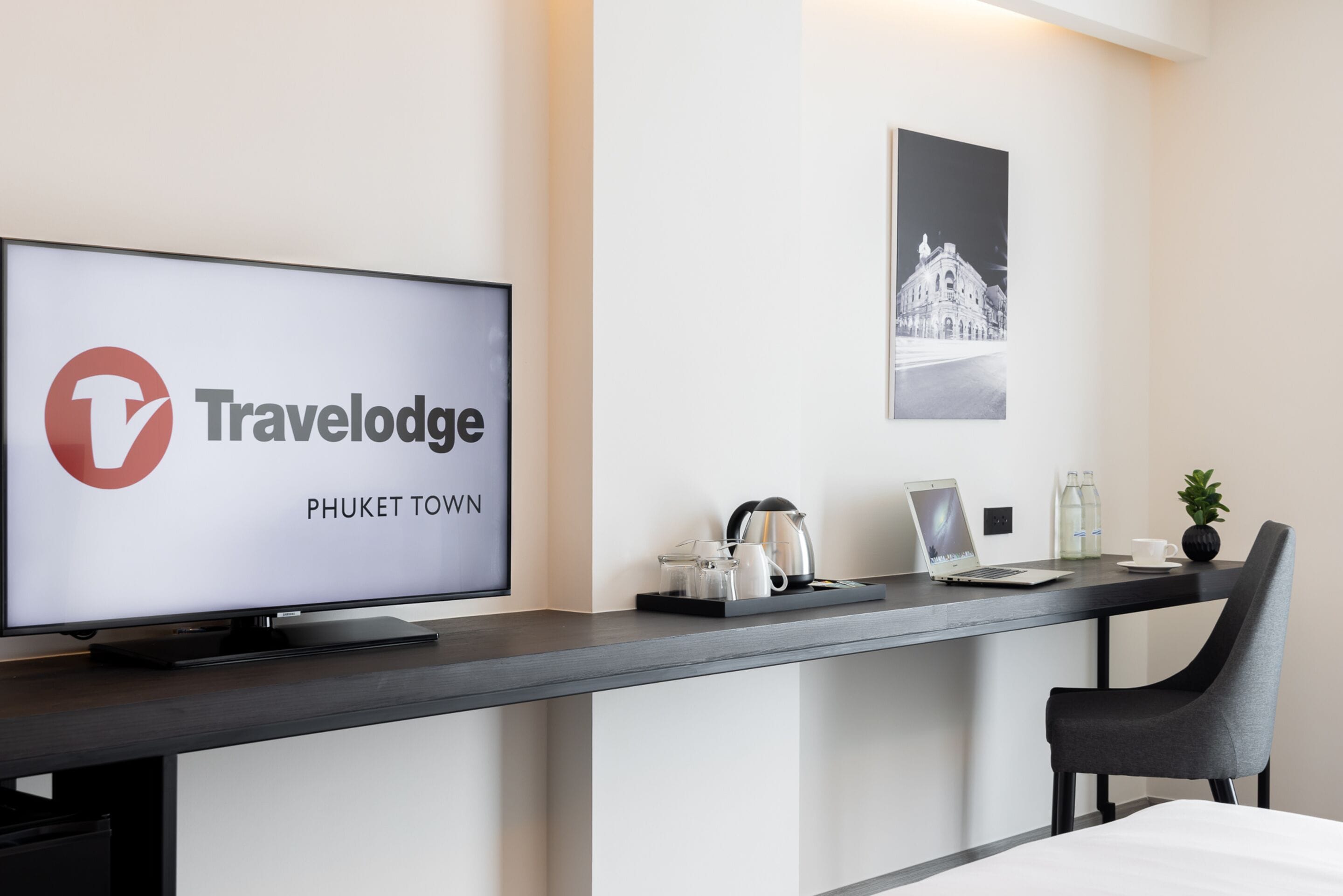 A modern hotel room with a large flat-screen TV displaying the Travelodge Phuket Town logo, a desk with a laptop, a kettle, and a small potted plant.