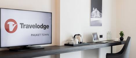 Una habitación de hotel moderna con una televisión de pantalla plana grande mostrando el logotipo de Travelodge Phuket Town, un escritorio con una computadora portátil, una tetera y una pequeña planta en maceta.