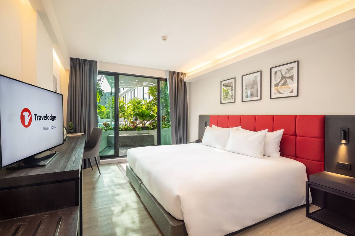 A modern hotel room with a large bed, a TV, a desk, and a balcony with plants.