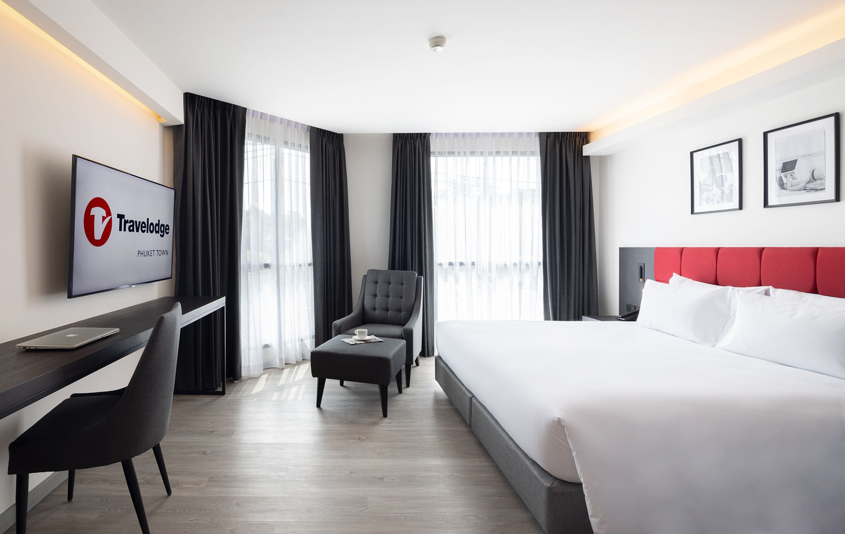 A hotel room with a bed, a desk with a laptop, a chair, a TV, and a large window with curtains.