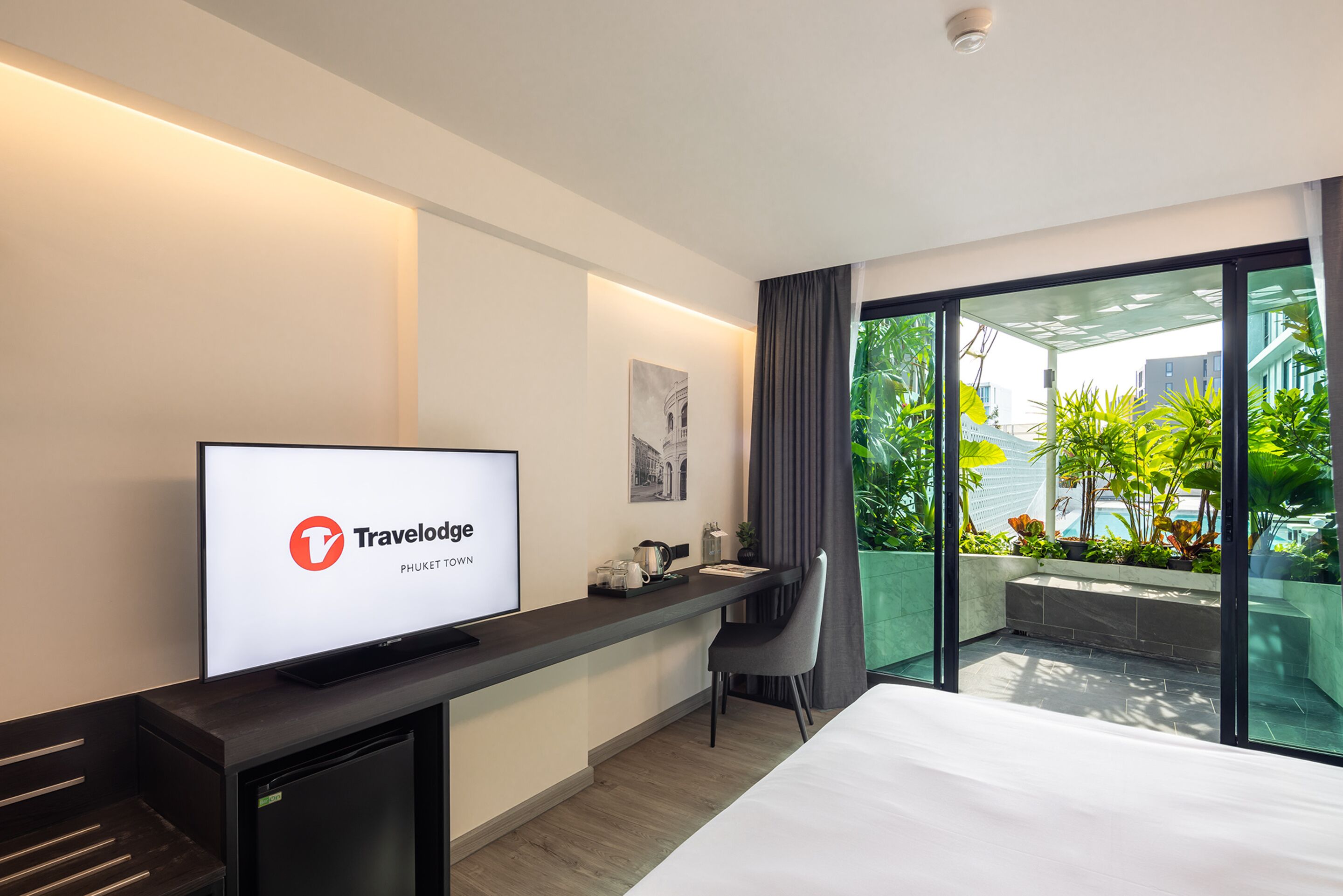 A modern hotel room with a large bed, a desk with a computer, a flat-screen TV displaying 'Travelodge Phuket Town', and a balcony with plants and a view of buildings.