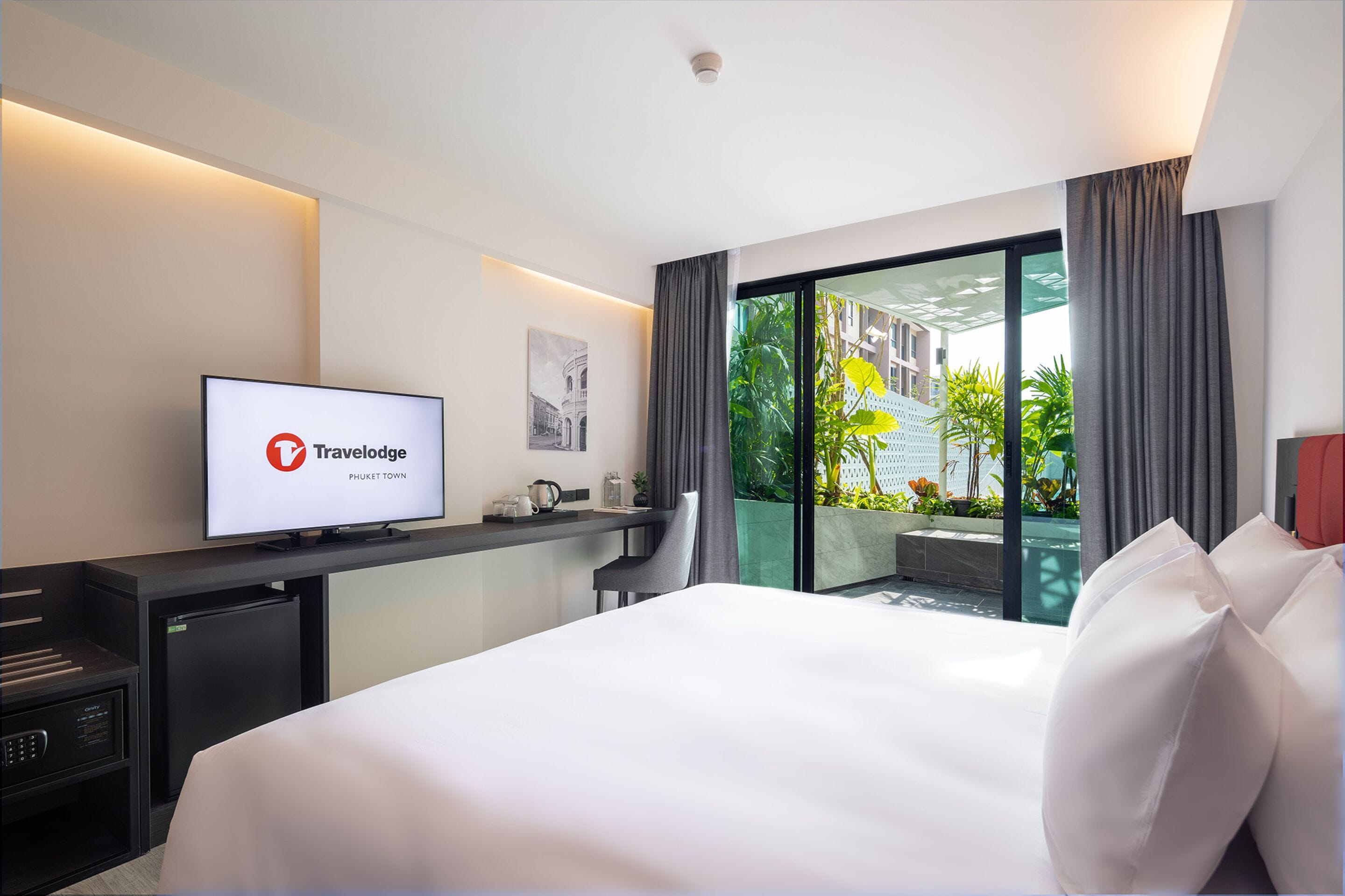 A modern hotel room with a bed, a desk with a computer, a television displaying 'Travelodge Phuket Town', and a view of greenery outside.