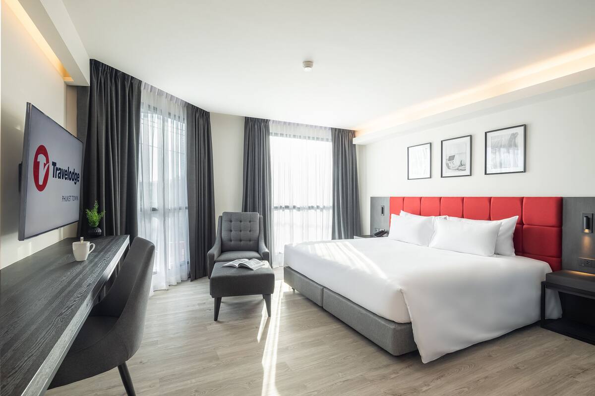 A modern hotel room with a large bed, a desk, a chair, and a TV displaying the Travelodge logo.