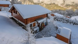Ein Holzchalet mit verschneitem Dach, einer kleinen Veranda und Blick auf verschneite Berge im Hintergrund.