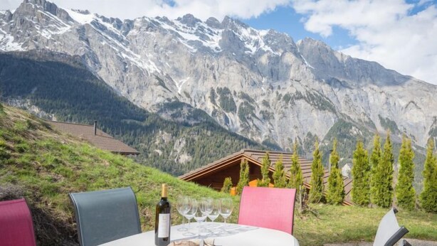 Ein runder Tisch mit einer Weinflasche, Gläsern, einem Brotlaib und einer Speisekarte, vor einer Berglandschaft mit einer Holzhütte und Kiefern im Hintergrund.