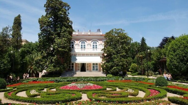 Un imposant bâtiment à la façade symétrique, entouré d’un jardin aménagé avec soin, présentant des motifs géométriques et de magnifiques parterres de fleurs.