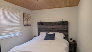 Ein Schlafzimmer mit einem Holzkopfende, einem Bett mit weißer Bettwäsche, einem Nachttisch, einer kleinen Topfpflanze und einem Fenster mit Jalousien.