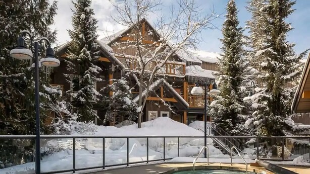 Un espace piscine doté d’une terrasse en bois, entouré d’arbres et de bâtiments couverts de neige.