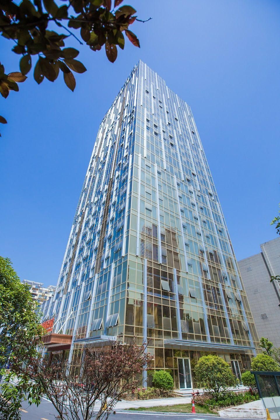 A tall, modern glass building with a grid-like facade, surrounded by greenery and other buildings.