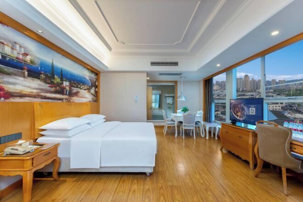 A hotel room with a bed, desk, chair, and a large window with a city view.