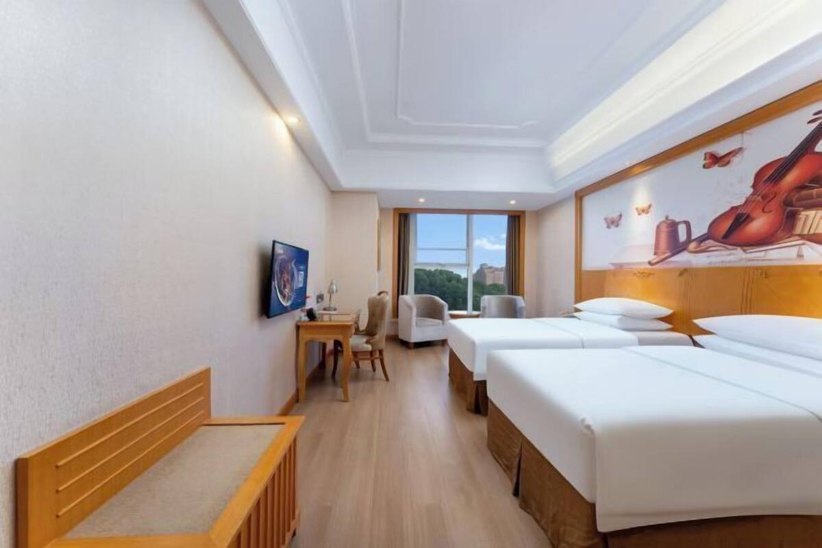 A hotel room with two beds, a desk, a chair, and a large window.