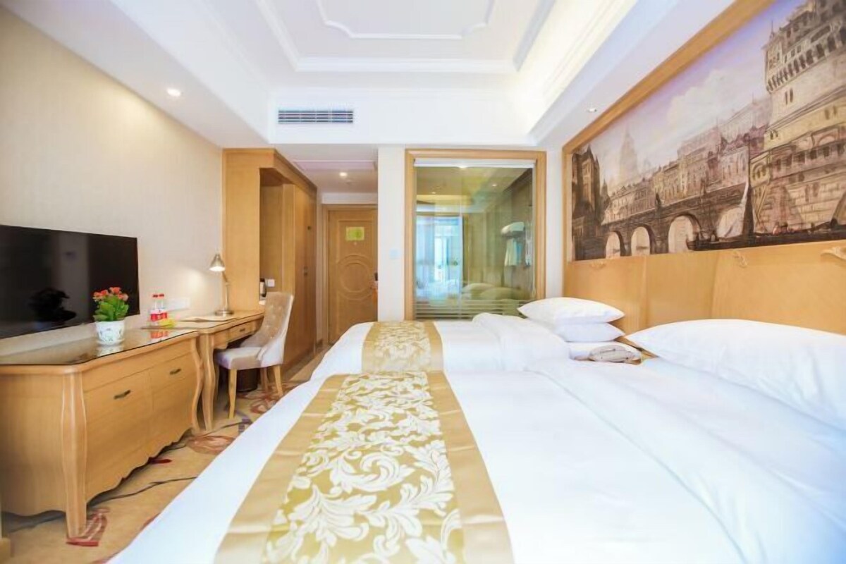 A hotel room with a bed, a desk with a chair, a television, and a painting of a bridge.