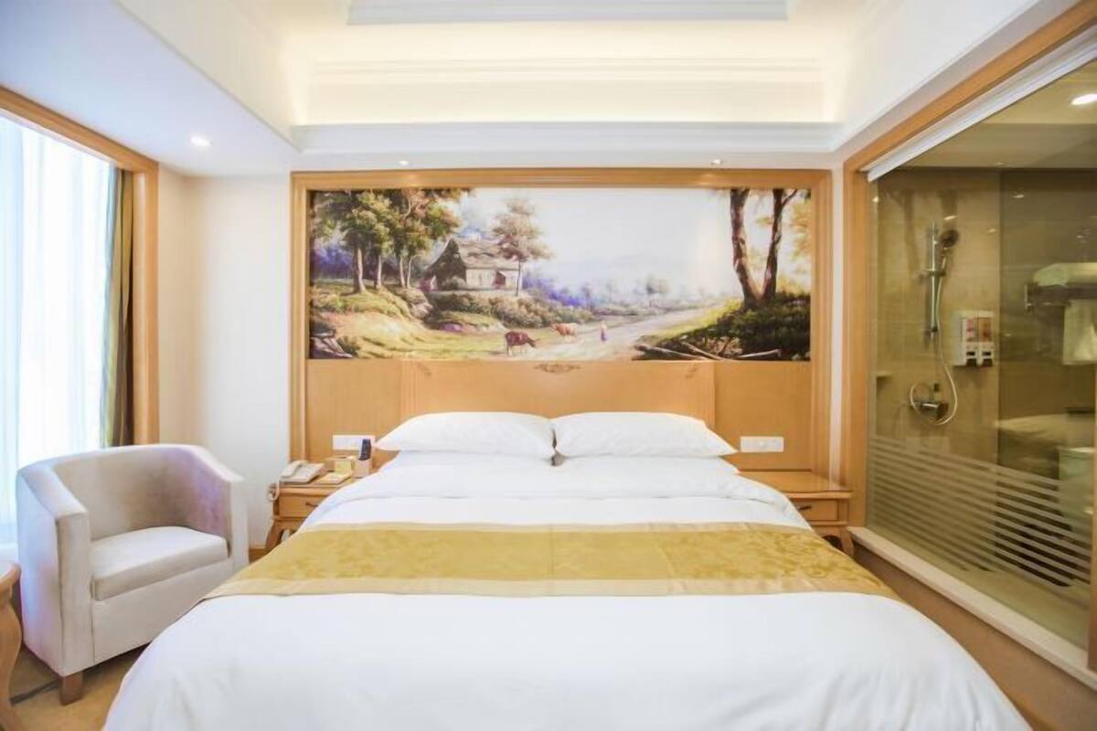 A hotel room with a large bed, a chair, a painting on the wall, and a glass shower enclosure.
