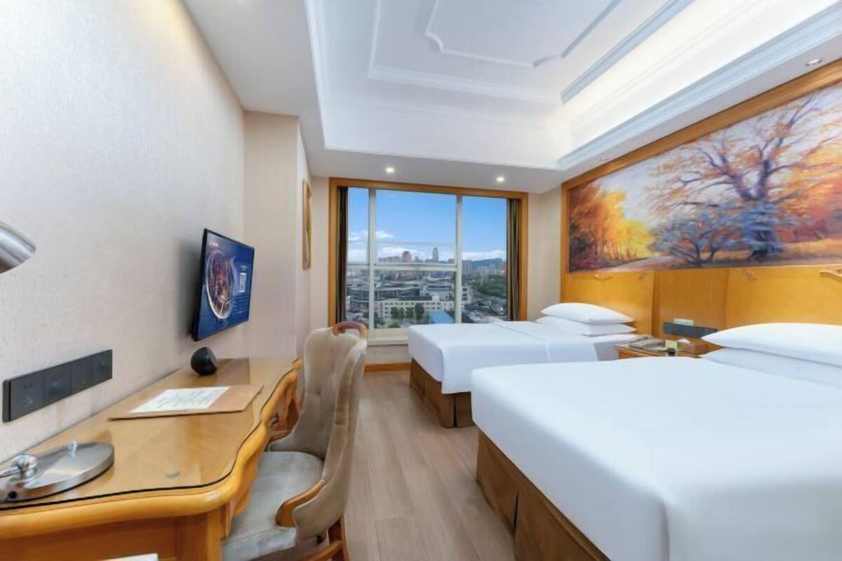 A hotel room with a desk, two beds, a chair, a painting, and a view of the city.