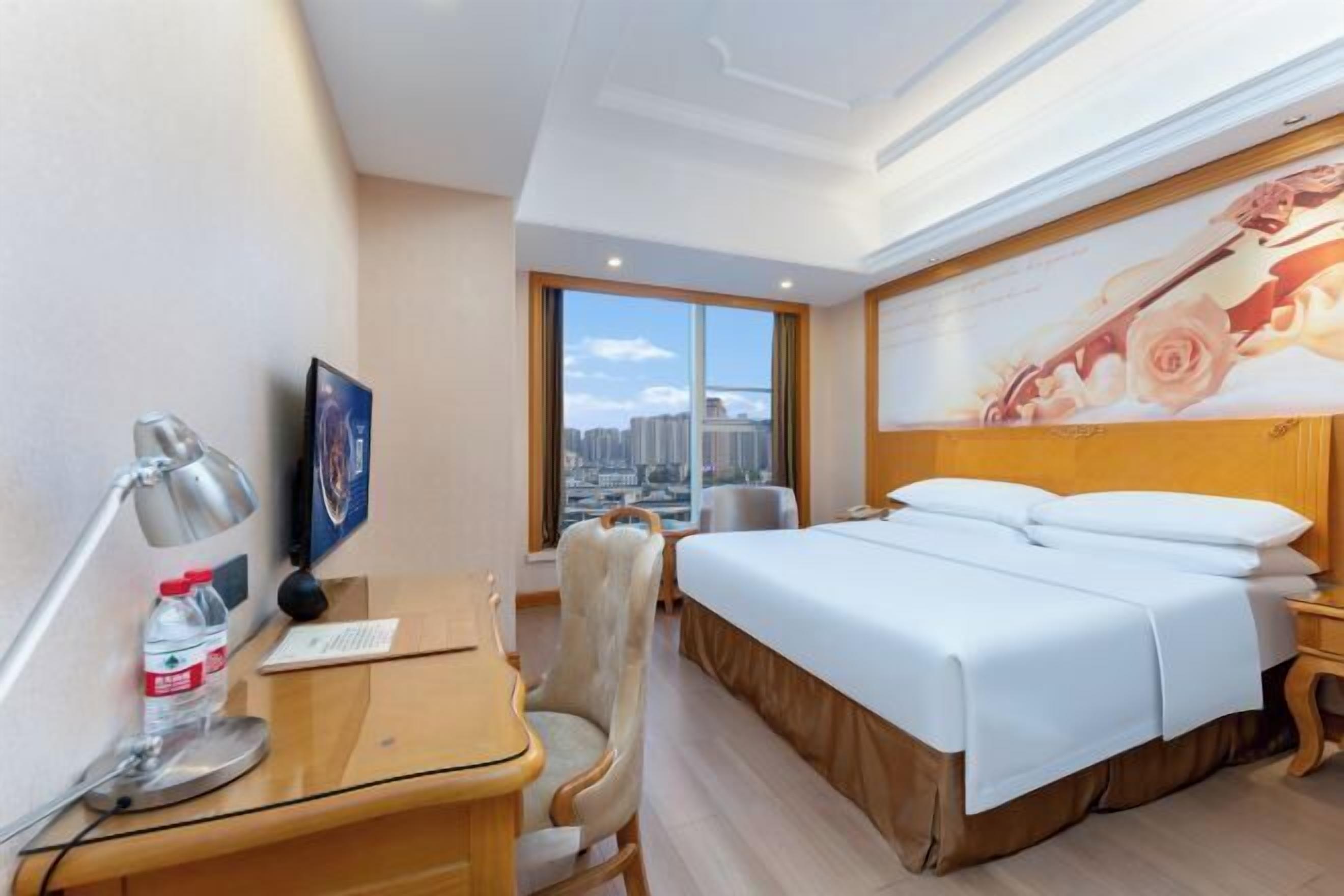 A hotel room with a bed, a desk with a lamp, a chair, a window with a city view, and a wall with a floral painting.