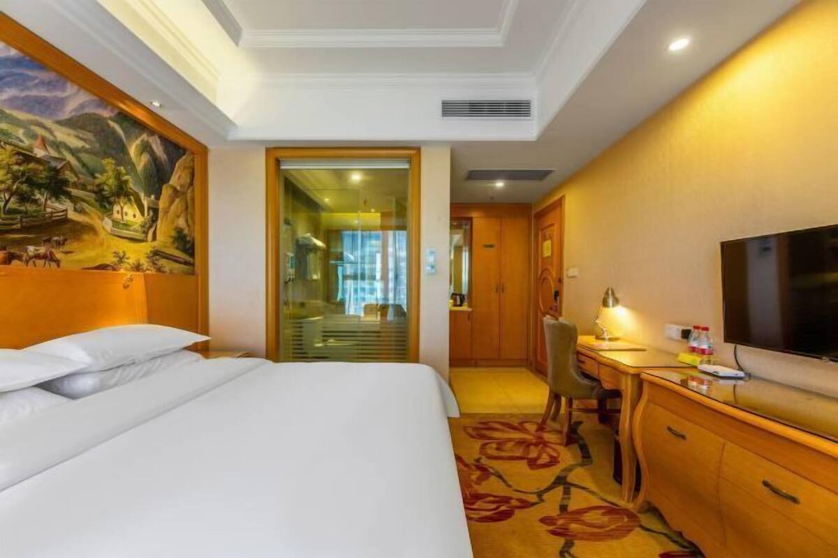 A hotel room with a bed, desk, TV, and a painting on the wall.
