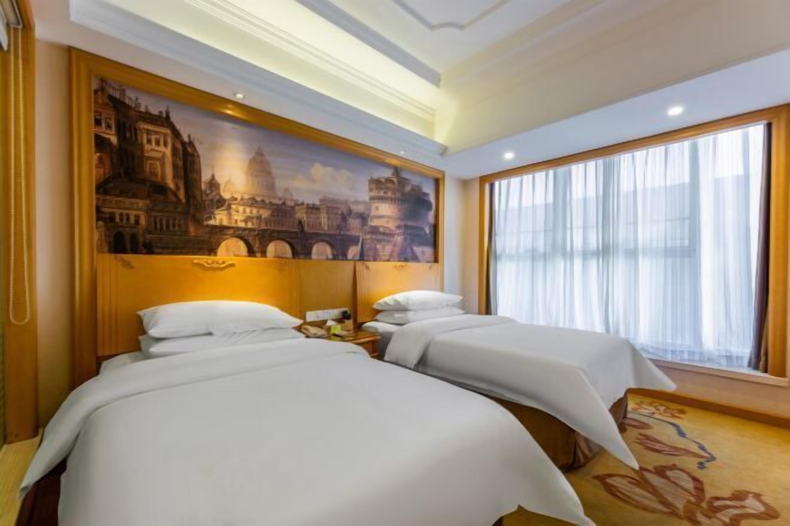 A hotel room with two beds, a large painting of a historical building, a nightstand with a lamp, and a large window with curtains.
