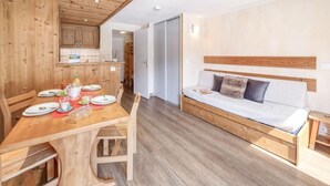 A modern, compact living space with a wooden dining table set for four, a sofa with a wooden headboard, and a kitchen area with wooden cabinets. - Palafour (Le Lac) by Interhome (Tignes)