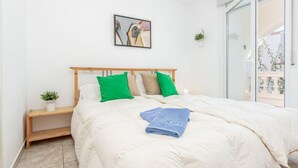 Un dormitorio con cama de madera, ropa de cama blanca, almohadas verdes, una toalla azul, una pequeña planta en la mesita de noche y un cuadro en la pared.