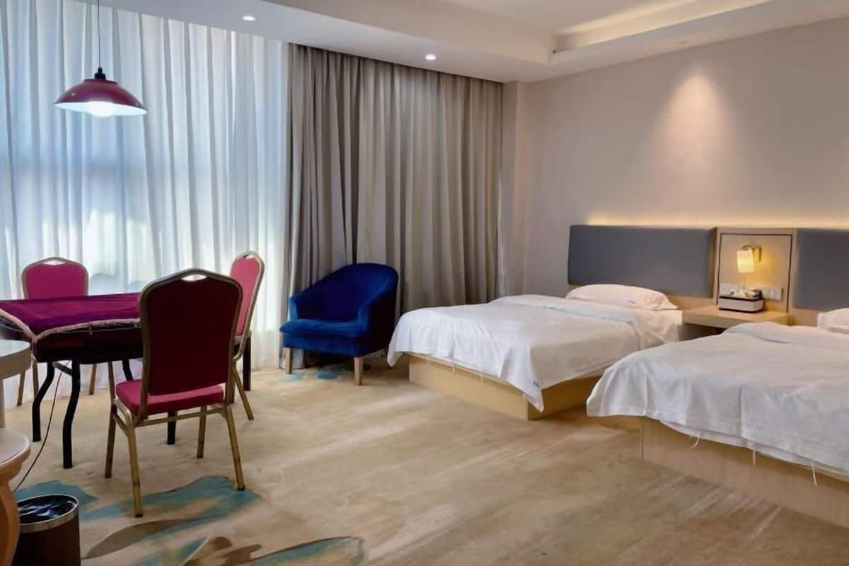 A hotel room with two beds, a dining table with chairs, a blue armchair, and a large window with curtains.