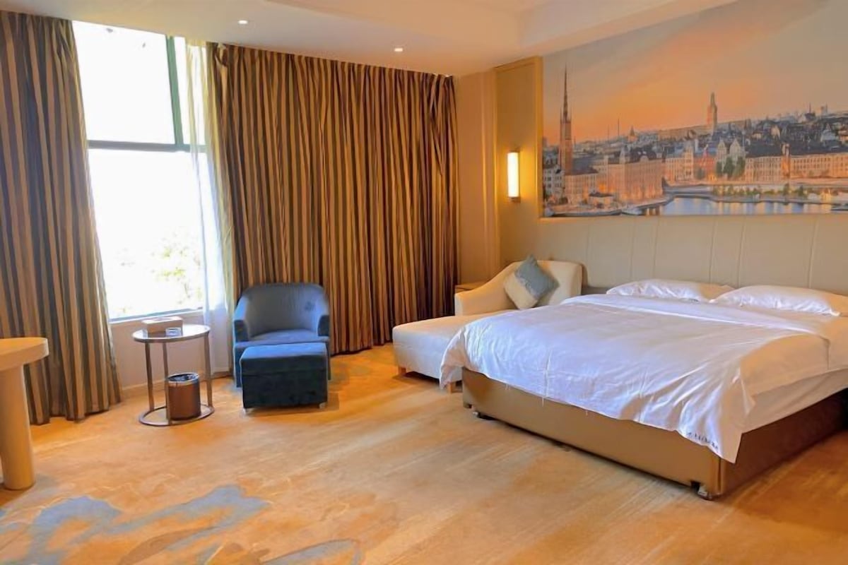 A hotel room with a large bed, a chair, a small table, and a painting of a cityscape on the wall.