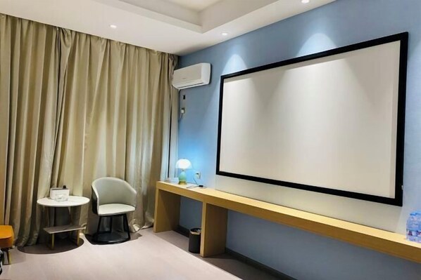 Habitación de hotel con una cama grande, un escritorio con silla, una mesita pequeña y una pantalla grande en la pared.
