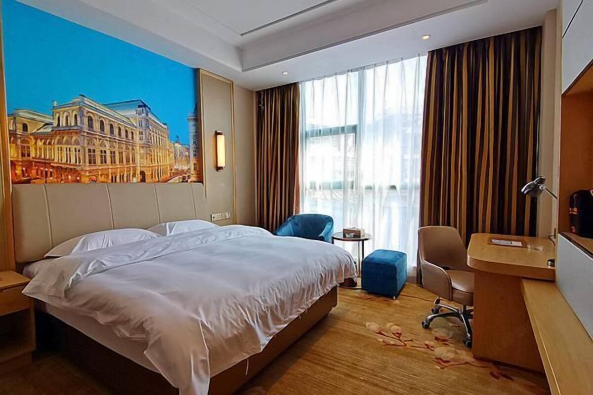 A hotel room with a large bed, a desk, a chair, and a view of a cityscape through the window.