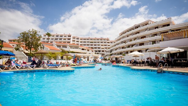 A large outdoor swimming pool surrounded by lounge chairs and umbrellas, with a multi-story hotel building in the background. - 2007 Garden Chic, Heated Pool (San Eugenio Alto)