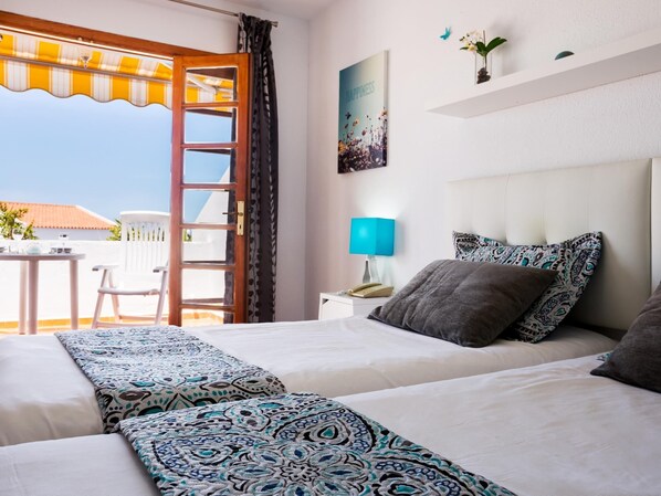 A hotel room with two beds, a balcony with a table and chairs, a nightstand with a lamp, and a wall with a framed picture. - 2007 Garden Chic, Heated Pool (San Eugenio Alto)