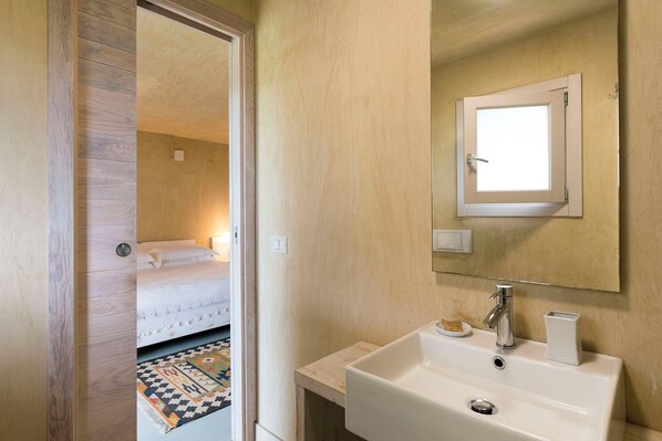 A hotel room with a wooden door, a bed with white bedding, a small bedside table with a lamp, a white sink with a mirror above it, and a patterned rug on the floor. - Converted farmhouse, nestled in nature, with pool (Caltagirone)
