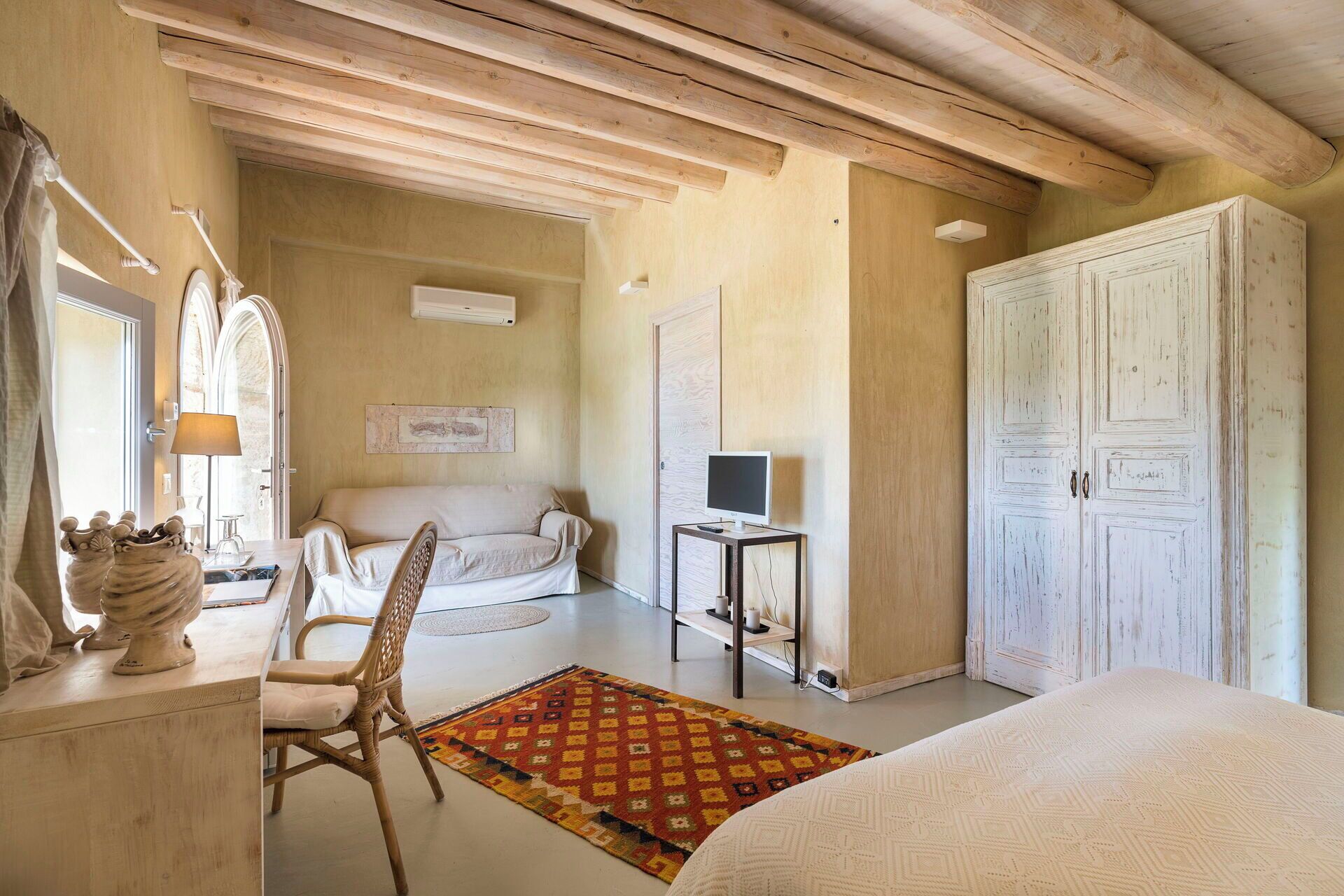 A bedroom with a bed, a chair, a desk with a lamp, a TV on a small table, and a wardrobe.