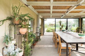An indoor dining area with a long table and chairs, surrounded by potted plants and a view of the garden outside. - Converted farmhouse, nestled in nature, with pool (Caltagirone)