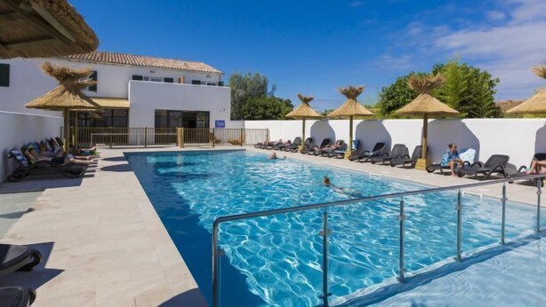 A swimming pool with lounge chairs and umbrellas, surrounded by a white building with a tiled roof. - Résidence Odalys Collection Rève d'Ile by Interhome (Ile de Re)