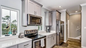 A modern kitchen with stainless steel appliances, white cabinetry, and a large window providing a view of the outdoors. - Closest Complex to Community Pool | Electric Car Charging Outlet | Private Beach Club | Golf |Tennis (Pensacola)