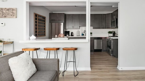 A modern kitchen with grey cabinets, a stainless steel refrigerator, and a white countertop. There are three wooden stools and a grey sofa with a white pillow. - Inviting Stylish Home with Covered Patio (Chicago)