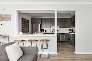 Een moderne keuken met grijze kastjes, een roestvrijstalen koelkast en een wit aanrecht. Drie houten krukken en een grijze bank met een wit kussen.