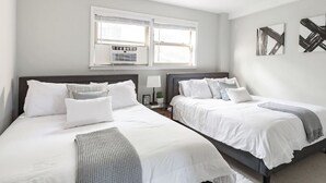 A modern bedroom with two beds, a large window with air conditioning, and abstract wall art. - Inviting Stylish Home with Covered Patio (Chicago)