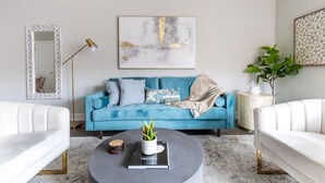 A modern living room with a blue sofa, a white armchair, a round coffee table with a book and a plant, a floor lamp, and a large abstract painting on the wall.
